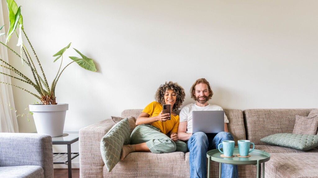 Couple assis sur un sofa, regardant leurs écrans en souriant - Taux fixe ou taux variable : comment choisir le bon taux hypothécaire? | Multi-Prêts Hypothèque