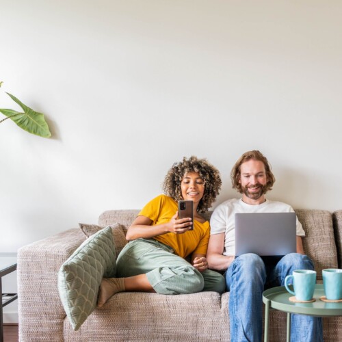 Couple assis sur un sofa, regardant leurs écrans en souriant - Taux fixe ou taux variable : comment choisir le bon taux hypothécaire? | Multi-Prêts Hypothèque